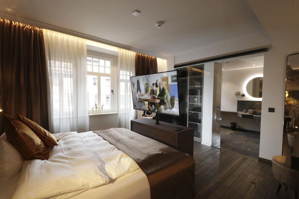 Blick auf das großzügige Doppelbett mit SmartTV und das Badezimmer mit gläserner Schiebetür.